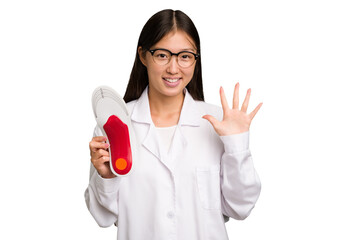 Wall Mural - Young asian chiropodist holding a insoles for shoes isolated smiling cheerful showing number five with fingers.