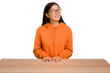 © Asier - Young asian woman sitting on a table isolated relaxed and happy laughing, neck stretched showing teeth.
