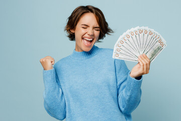 Wall Mural - Young happy caucasian woman wear knitted sweater look camera hold in hand fan of cash money in dollar banknotes do winner gesture isolated on plain pastel light blue cyan background studio portrait.