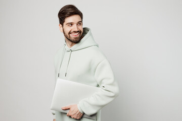 Young fun programmer happy caucasian man wear mint hoody hold closed laptop pc computer look aside on workspace area isolated on plain solid white background studio portrait. People lifestyle concept.