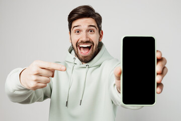 Young amazed fun impressed happy man wear mint hoody hold in hand use point index finger on mobile cell phone with blank screen workspace area isolated on plain solid white background studio portrait.