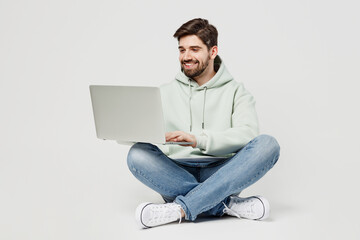 Full body smiling happy programmer smart young caucasian IT man wear mint hoody hold use work on laptop pc computer isolated on plain solid white background studio portrait. People lifestyle concept.
