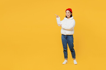 Wall Mural - Full body happy smiling young woman of Asian ethnicity wear white padded windbreaker jacket red hat pointing indicate on workspace area copy space mock up isolated on plain yellow background studio.