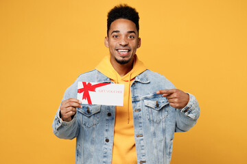 Sticker - Young surprised amazed man of African American ethnicity wear denim jacket hoody hold point index finger on gift certificate coupon voucher card for store isolated on plain yellow background studio.