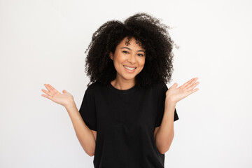 Wall Mural - Cheerful young woman shrugging throwing up hands. Hispanic female model with afro hairstyle and brown eyes in casual clothes smiling, spreading her hands looking confused. Uncertainty, doubt concept