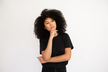 Wall Mural - Pensive young woman looking away holding thumb on her chin. Hispanic female model with afro hairstyle and brown eyes in black T-shirt looking away touching chin with thumb. Thought, dream concept