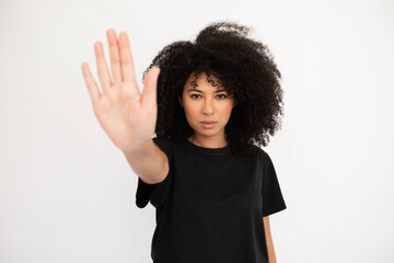 Wall Mural - Serious young woman showing stop sign. Caucasian female model with afro hairstyle and brown eyes in black T-shirt showing her palm as stop sign. Forbiddance, advertising concept