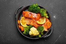 Healthy Baked Fish Salmon Steaks, Broccoli, Cauliflower, Carrot In Cast Iron Casserole Bowl Black Dark Stone Background. Cooking A Delicious Low Carb Dinner, Healthy Nutrition