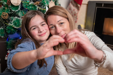 Loving family spends winter holidays at home. Portrait of happy smiling beautiful mother and child joining hands making heart shape sitting in festive, cozy room with Christmas tree .