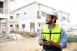 © bigy9950 - portrait of young engineer in vest with white helmet standing on construction site, smiling and holding smartphone for worker, internet, social media.
