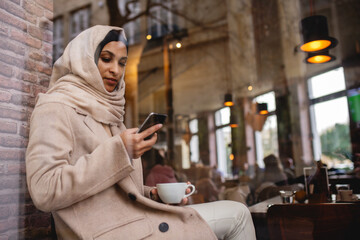 Sticker - Young muslim woman with smartphone enjoying cup of coffe in cafe.