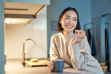 Beautiful young asian woman eating toast on her kitchen, drinking coffee and smiling