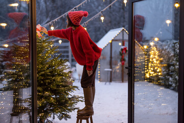 Young woman in red decorates lush Christmas tree with festive ballls and garland at backyard of her house on snow fall, preparing for a winter holidays