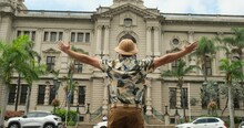 Man In Safari Clothes Raised His Hands Against The Background Of An Old Building With The Flag Of South Africa. American Male Traveler Sit And Relax In The Park And During Vacation Enjoys Architecture