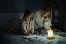 Mother And Little Daughter Studying And Drawing In A Complete Darkness During Electricity Outage. Little Girl Uses Camping Lantern To Do Her Homework During Blackout. Energy Crisis Concept