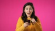 © Robert Peak - Portrait of happy smiling young pretty woman 20s in sweater shopping on mobile phone point with finger give thumbs up isolated on pink background studio.