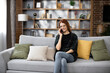 © sofiko14 - Happy young caucasian woman sitting on cozy sofa with modern smartphone in hands and calling to customers during break. Pretty female resting during working day at home.