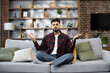 © sofiko14 - Calm of bearded young man doing yoga lotus pose to meditation and relax on couch during work online at home. Happy male close his eyes after hard work during break and deep breath in yoga posotion
