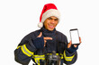 © Serhii - Young smiling African-American fireman in uniform and red Santa Hat holding smartphone in hand indicating, finger advising discount price looking at camera isolated on white background, copy space
