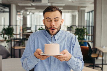 Excited man celebrating birthday in office, looking happy, holding cake with candle, making a wish.