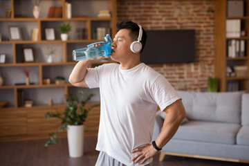 Wall Mural - Sporty mature korean man with headphones drinking water after workout, staying hydrated during domestic sports training