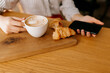 Flat lay of cappuccino with croissant on wood table. Woman hand is holding cup of coffee and smartphone 