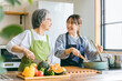 家のキッチンで一緒に料理・家事・自炊をする笑顔のアジア人高齢者女性と若い女性（家政婦・家事代行）
