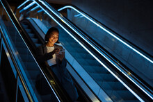 Young Asian Woman Using Mobile Phone While Standing On Escalator Indoors