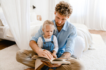 Young white father and little daughter reading book together at home