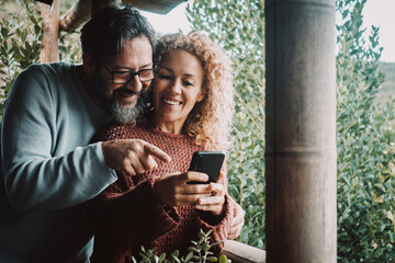 Happy couple using mobile phone having fun together outside on the terrace at home. Man and woman smile and use cellular to video call and communicate in outdoor. Technology and people lifestyle