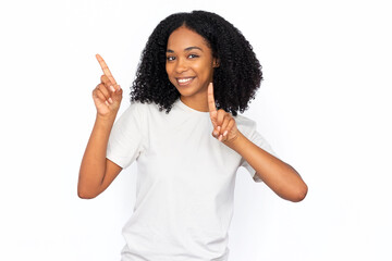 Wall Mural - Excited African American woman pointing up. Portrait of happy young female model with dark curly hair in white T-shirt looking at camera, smiling, showing ads with fingers. Advertisement concept