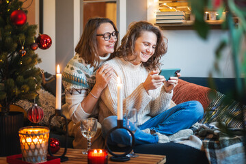 Smiling embracing happy lesbian couple in warm sweaters sitting on sofa at home near christmas tree and using smartphone