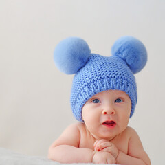 Wall Mural - A happy newborn baby in a blue hat with pompoms looks into the camera. A smiling two-month-old infant is lying on a blanket. Beautiful, happy little child with blue eyes.  Cheerful White kid. .