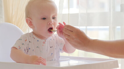 Wall Mural - Mother is Feeding a baby from a spoon. Mom feeds a 9 month baby with fruit puree from a spoon, close-up, high key. Infant boy eats sitting on baby's chair. Mother cares about little son.