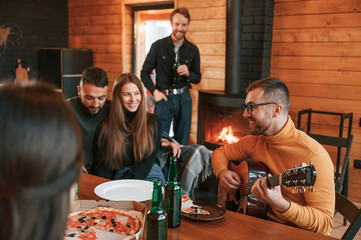 Wall Mural - Man in glasses is playing guitar. Group of friends is having good weekend indoors in the wooden building together