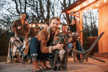 Wall Mural - Happy man is sitting with his dog. Group of people is spending time together on the backyard at evening time