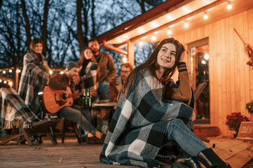 Wall Mural - Young woman with a blanket is sitting. Group of people is spending time together on the backyard at evening time