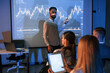 © standret - People are sitting. Male leader talking to employees, showing the plan on the projector in office of stock exchange company