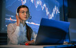 © standret - Sitting by laptop. Female stock broker is working indoors. Big wide display behind her