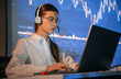 © standret - Sitting by laptop. Female stock broker is working indoors. Big wide display behind her