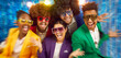 Portrait of laughing happy people in disco style sunglasses, colourful suits on blue blur background looking at camera. Group of friends in funny curly wigs having fun at night club on disco party.