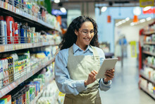 Latin American Woman Seller Happy And Smiling Uses Tablet Computer In Supermarket To Check Stock Availability, Shop Assistant Among Shelves With Goods In Apron.