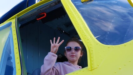 Child girl sits in cockpit of yellow helicopter and shows sign V with fingers. Girl 10-15 years old in sunglasses and dusty pink sweatshirt sits in cockpit of helicopter and smiles, showing V gesture.