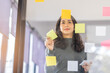 © David - Business female employee with many conflicting priorities arranging sticky notes commenting and brainstorming on work priorities colleague in a modern office.