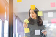 © David - Business female employee with many conflicting priorities arranging sticky notes commenting and brainstorming on work priorities colleague in a modern co-working space.
