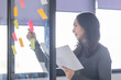 © David - Business female employee with many conflicting priorities arranging sticky notes commenting and brainstorming on work priorities colleague in a modern co-working space.