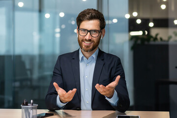 Portrait of a friendly young handsome man sitting in the office at the table wearing headphones and talking to the camera. Business meeting, tutor support, job interview, consultation