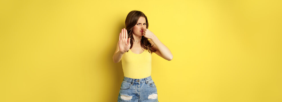 Woman showing stop sign and shut nose from disgust, dislike bad smell, declining something awful, standing over yellow background