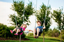 Young Girl And Boy Holding Hands While Playing On A Swing Set At A Playground; St. Albert, Alberta, Canada