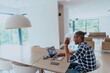 © .shock - African American man in glasses sitting at a table in a modern living room, using a laptop for business video chat, conversation with friends and entertainment